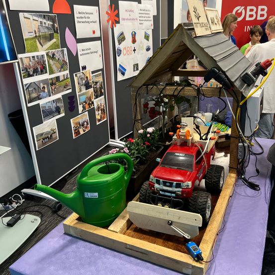 Eine Holzstruktur ähnelt einem Haus mit Dach und beherbergt einen roten Spielzeug-LKW im Inneren. Ein grüner Gießkanister ist auf der linken Seite platziert, und verschiedene elektronische Komponenten und Drähte sind angeschlossen. Im Hintergrund sind Menschen zu sehen.