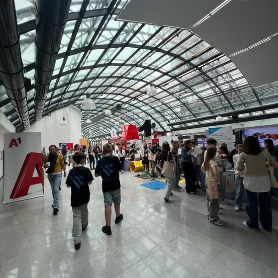 Ein überfüllter Innenbereich mit Glasdach, in dem Menschen herumlaufen, einige mit Taschen. Ein großes rotes 'A' befindet sich links und ein Bildschirm rechts. Die Leute sind um die Stände versammelt.