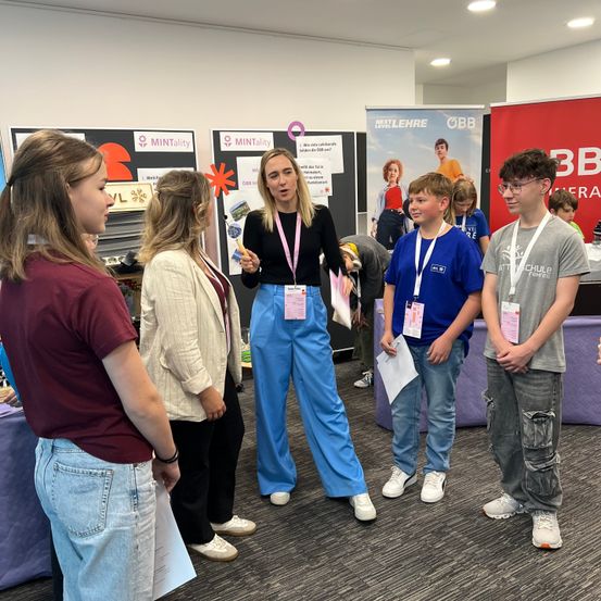 Eine Gruppe junger Menschen steht in einem Raum, eine Frau spricht zu ihnen. Die Frau trägt ein schwarzes Oberteil und blaue Hosen. Die Gruppe trägt Namensschilder. Hinter ihnen sind Poster mit dem Text MINTailty und VL.