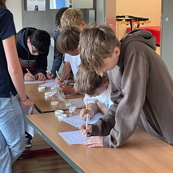 Schüler arbeiten an einem Tisch mit Papieren, Stiften und kleinen Gläsern und sind an einem Experiment beteiligt. Ein Schüler beugt sich mit einem Stift in der Hand über den Tisch.