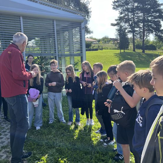 Eine Gruppe von Schülern und ihrem Lehrer versammelt sich auf einer Wiese, einige halten Gegenstände, andere schauen zu, mit einem Gebäude und Bäumen im Hintergrund.