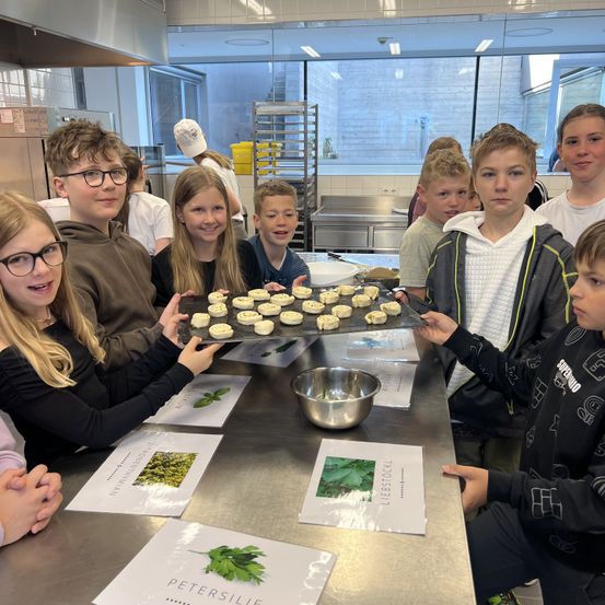 Eine Gruppe junger Menschen in einer Küche, die ein Tablett mit Keksen halten und lächeln. Sie haben Rezeptpapiere vor sich.