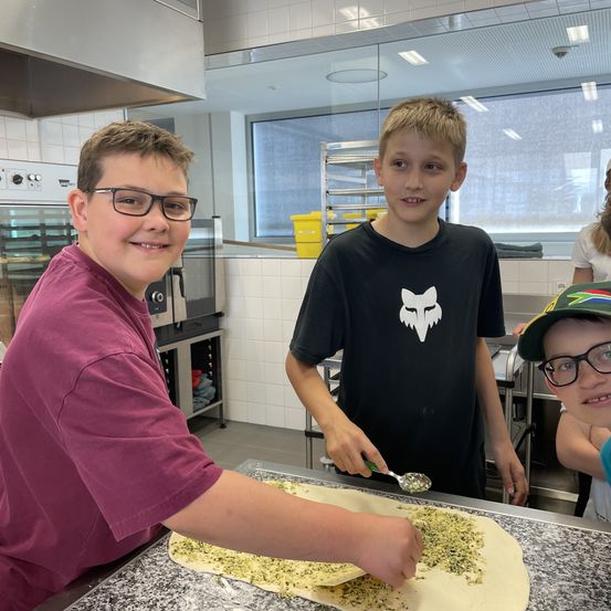 Drei Jungen in einer Küche beim Pizzamachen. Einer verteilt Pesto, ein anderer hält einen Löffel und der dritte beobachtet.