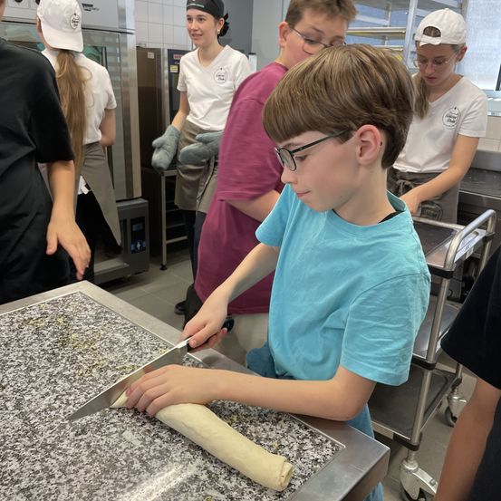 Eine Gruppe von Schülern befindet sich in einer Küche, wobei ein Junge Teig mit einem Messer schneidet. Hinter ihm beobachtet ein Mädchen. Andere sind ebenfalls in der Nähe und arbeiten an Teig.