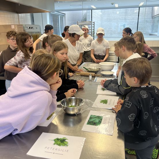 Eine Gruppe von Kindern und Erwachsenen in einer Küche, einige in weißen Hemden und Mützen, betrachten einen Tisch mit Papieren und Schüsseln. Eine Frau in einer weißen Mütze steht vor ihnen.