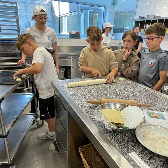In einer Küche arbeiten mehrere Kinder an Teig, angeleitet von einem Erwachsenen. Schüsseln, ein Nudelholz und Tabletts mit Teig stehen auf dem Tresen.