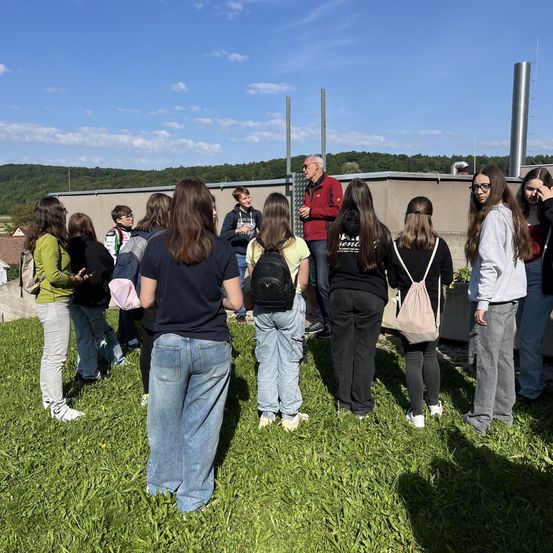 Eine Gruppe von Schülern hört einem Mann zu, der in der Nähe eines Metallbaues steht, an einem sonnigen Tag. Sie stehen auf einem Rasenbereich vor einem Gebäude.