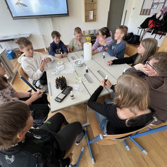 Eine Gruppe von Kindern sitzt um einen Tisch herum mit verschiedenen Gegenständen, möglicherweise in der Durchführung eines wissenschaftlichen Experiments. Sie sind in Aktivitäten mit Flüssigkeiten und kleinen Behältern engagiert. Eine Projektionsleinwand ist an der Wand hinter ihnen angebracht.