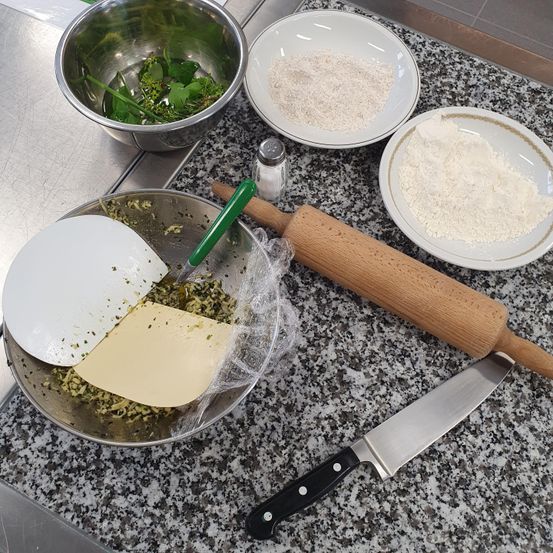 Ein Küchentisch zeigt eine Schüssel mit gehackten Kräutern, eine Schüssel mit Mehl und eine Schüssel mit einem Nudelholz. Ein Messer und eine zerbrochene Pfanne sind ebenfalls vorhanden.