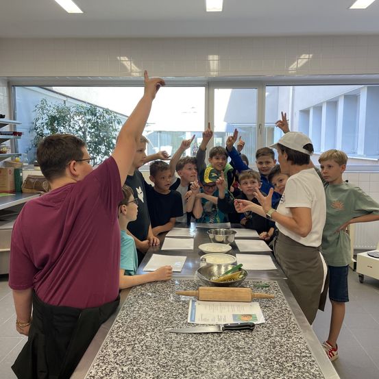 Eine Gruppe von Kindern in einer Küche mit einer Frau, die Gesten macht. Sie machen Friedenszeichen und schauen sie an.
