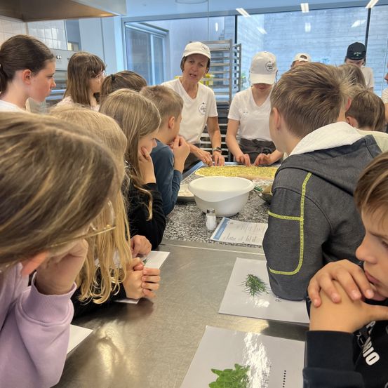Eine Gruppe von Kindern und zwei Frauen bereitet in einer Küche Essen zu. Eine Frau spricht, während die andere an einem Tisch mit Essen arbeitet.