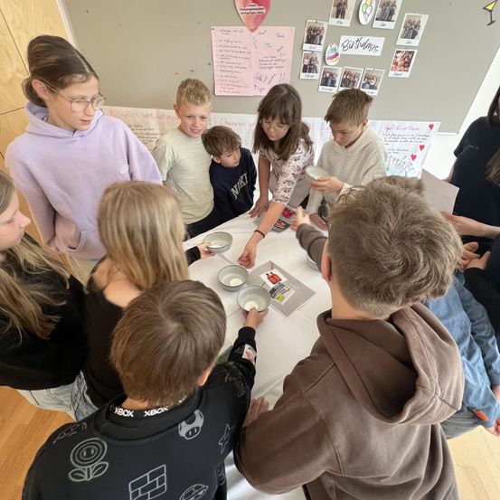 Eine Gruppe von Kindern und Erwachsenen versammelt sich um einen Tisch mit Schüsseln und Papieren, vielleicht in einer Klassenzimmersituation. Sie sind umgeben von einer Pinnwand mit verschiedenen Bildern und Schriften.