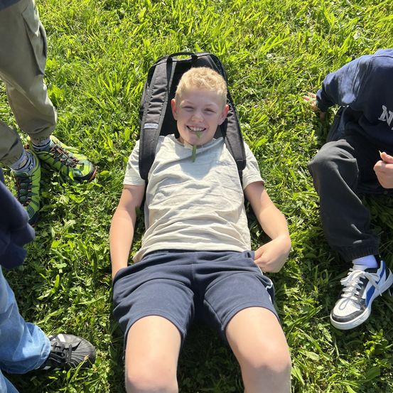 Ein Junge mit Rucksack liegt auf dem Gras und lächelt, während er eine Pflanze im Mund hält. Zwei andere Kinder in Turnschuhen sind in der Nähe.