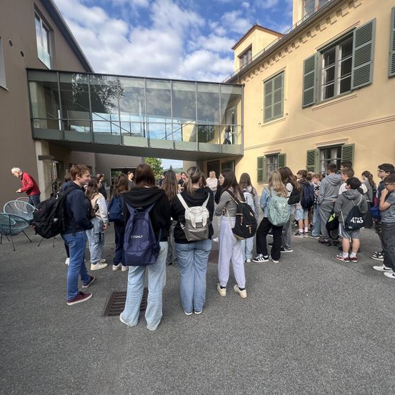Eine Gruppe von Schülern mit Rucksäcken wartet vor einem Gebäude mit Glasfenstern und einer Überführung.