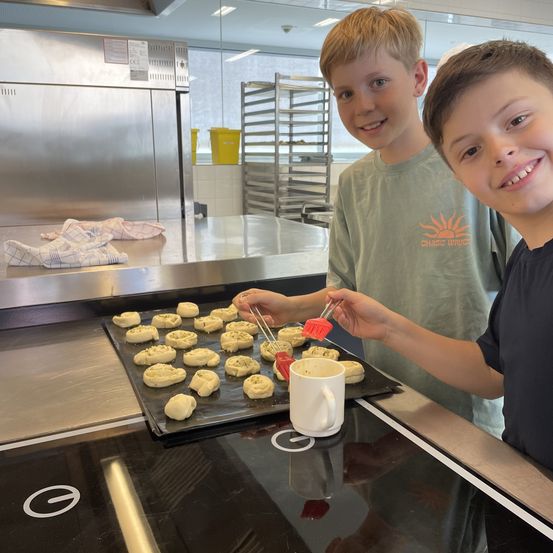Zwei junge Jungen sind in einer Küche, einer lächelt in die Kamera, der andere gibt den auf einem Backblech liegenden Teigwaren Toppings. Die Küche ist mit Edelstahlgeräten gut ausgestattet.