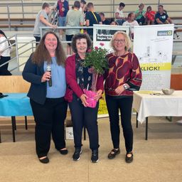 Drei Frauen stehen in einem Raum mit einem Podium, eine hält ein Mikrofon, eine andere Blumen und die dritte trägt eine Uhr. Dahinter stehen Tische mit weißen Tüchern.