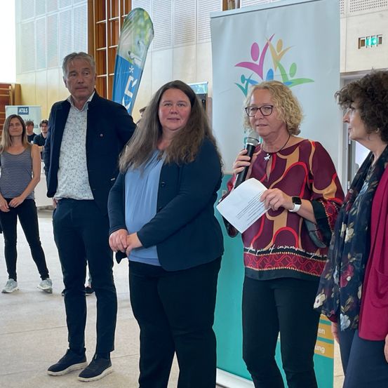 Eine Frau spricht in ein Mikrofon und hält ein Papier in der Hand. Hinter ihr hört eine Frau mit Brille zu. Daneben stehen ein Mann und eine weitere Frau. Hinter den Sprechern schaut eine Frau zu. Im Hintergrund sind Banner angebracht.