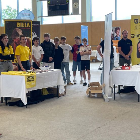 Eine Gruppe junger Menschen in verschiedenen Trikots steht hinter einem Tisch mit einem gelben Banner. Sie scheinen sich auf eine Veranstaltung vorzubereiten. Ein Mann steht hinter einem Banner mit einem Bild einer Frau.