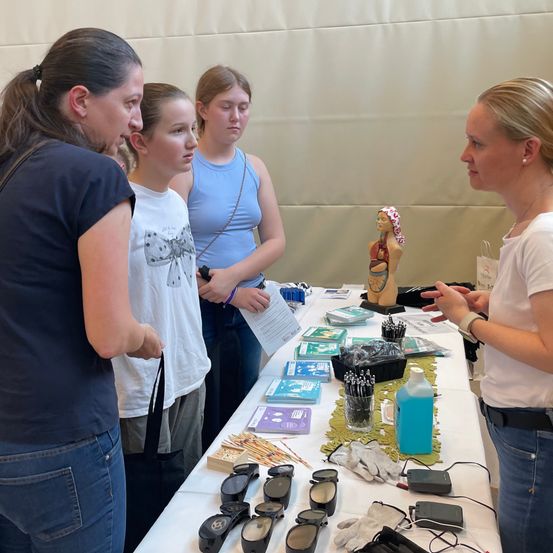 Vier Personen, darunter drei Mädchen und eine Frau, stehen um einen Tisch mit verschiedenen Gegenständen, möglicherweise bei einer Veranstaltung. Die Frau führt ein Gespräch mit den Mädchen.