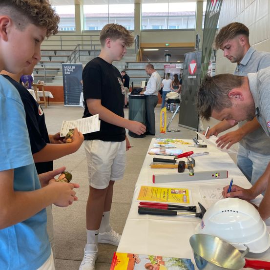 Eine Gruppe junger Männer auf einer Baumesse, zwei unterschreiben Papiere und zwei halten Spielzeug.