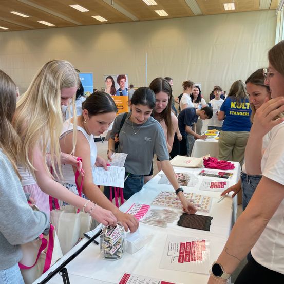 Mehrere Frauen versammeln sich um einen Tisch, möglicherweise auf einem Jahrmarkt, mit Gegenständen auf dem Tisch, darunter Bonbons und Papiere. Eine Frau hält ein Stück Papier und schaut sich die Gegenstände an. Eine andere Frau hält einen Stift und schreibt auf ein Stück Papier.
