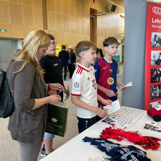 Eine Frau gibt einem Jungen mit Fußballtrikot und zwei anderen Jungen Gegenstände aus, in der Nähe eines Tisches mit Merchandise, in einem großen Raum.