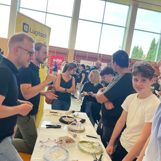 Mehrere Personen versammeln sich um einen Tisch mit verschiedenen Gegenständen. Ein Mann kniet und gestikuliert, während er mit einem anderen Mann spricht. In der Nähe hält eine Frau eine Tasche. Der Hintergrund besteht aus großen Fenstern mit Bannern auf beiden Seiten.