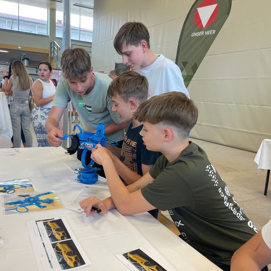 Vier junge Jungen sind um einen Tisch versammelt und konzentrieren sich auf ein blaues mechanisches Modell. Sie sind von gedruckten Bildern und einer weißen Tischdecke umgeben. Dahinter ist ein weißer Banner mit Text zu sehen, und mehrere Personen stehen im Hintergrund.