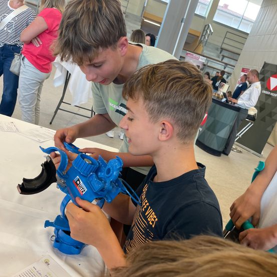 Zwei junge Jungen arbeiten gemeinsam an einem blauen mechanischen Objekt an einem Tisch, umgeben von einem geschäftigen Raum.