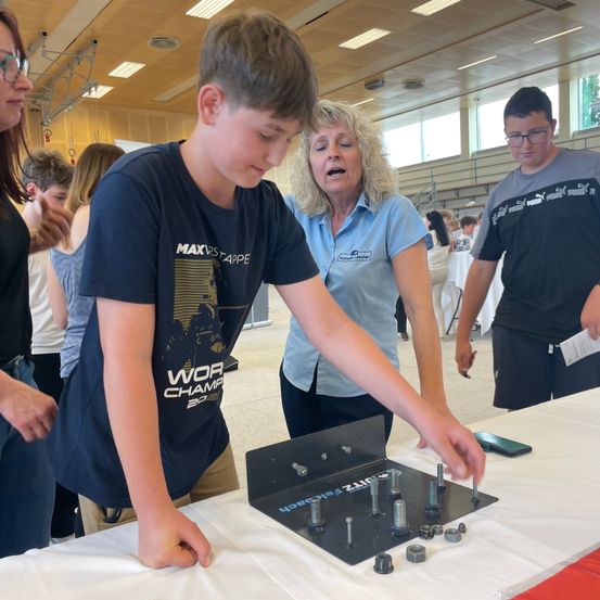 Ein junger Junge arbeitet an einem Projekt am Tisch, angeleitet von einer Frau. Er ist von anderen Menschen umgeben, einige stehend und einige sitzend. Der Tisch ist mit verschiedenen Werkzeugen und Komponenten bestückt.