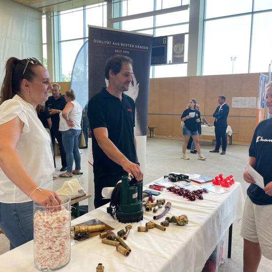 Ein Mann und eine Frau stehen an einem Tisch mit verschiedenen Gegenständen. Hinter ihnen unterhalten sich andere. Der Raum hat große Fenster und ein Banner mit dem Text 'Qualität aus besten Händen seit 1976'.