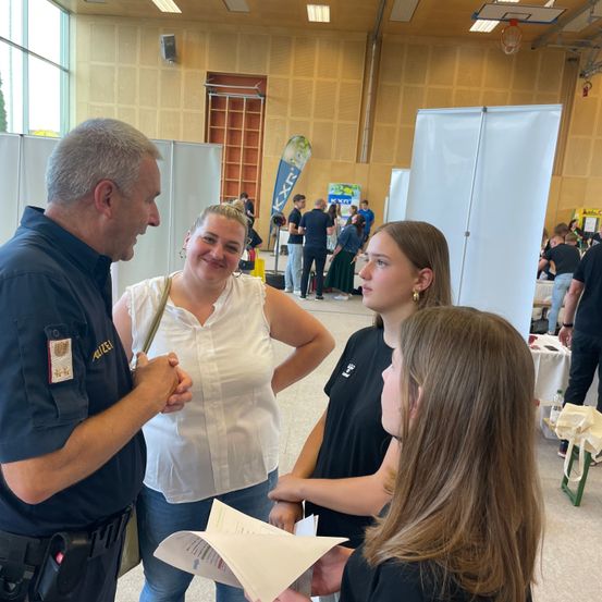 Ein Mann in einer Polizeiuniform spricht mit zwei Frauen und einem Mädchen. Die Frau hält ein Stück Papier, und sie stehen in einem Saal mit weißen Wänden.