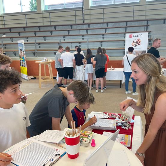 Eine Gruppe von Menschen, darunter Kinder, befindet sich bei einer Veranstaltung in einer Halle mit leeren Tribünen. Eine Frau spricht mit drei Jungen, die sich Gegenstände auf einem Tisch ansehen.