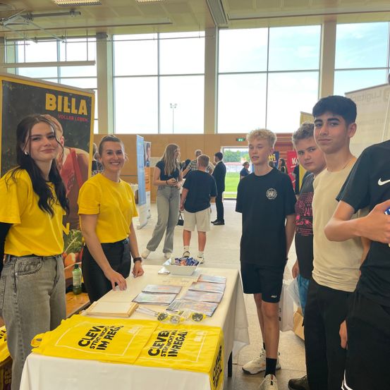 Mehrere Personen versammeln sich um einen Tisch mit Werbematerialien. Zwei Frauen in gelben T-Shirts stehen neben dem Tisch. Ein paar Jungen stehen rechts vom Tisch. Ein großes Plakat mit dem Text 'BILLA' ist an der Wand hinter ihnen befestigt.