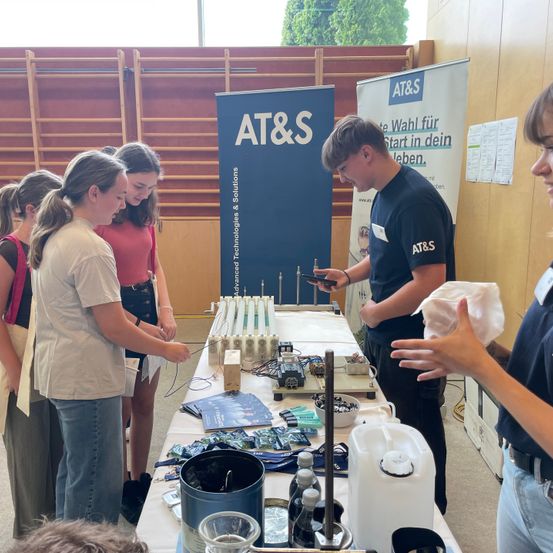 Eine Gruppe von Menschen versammelt sich um einen Tisch mit elektronischen Komponenten, ein Mann demonstriert etwas auf seinem Handy. Dahinter steht ein Hintergrund mit dem AT&S-Logo.