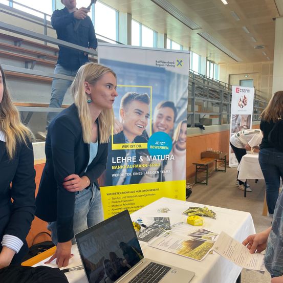 Mehrere Frauen stehen um einen Tisch mit Laptop und Broschüren. Ein Schild dahinter bewirbt Lehrstellen und Jobs. Eine Person steht auf der Tribüne darüber.