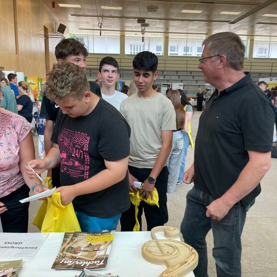 Eine Gruppe von Schülern, einige mit Taschen, steht in einer Schlange. Ein Mann signiert etwas. Es gibt Broschüren auf einem Tisch und ein Musiknoten-Ausschnitt.