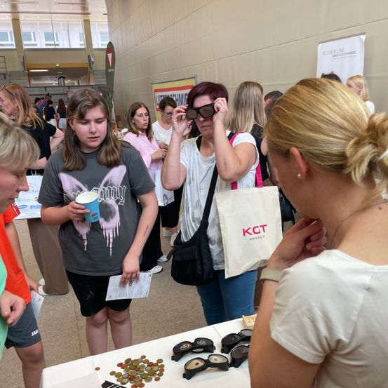Mehrere Personen, darunter junge Mädchen und Frauen, stehen um einen Tisch mit Sonnenbrillen. Eine Frau probiert eine Sonnenbrille an und eine andere hält einen Becher. Auf dem Tisch liegen Münzen und eine Tasche mit den Buchstaben KGT.