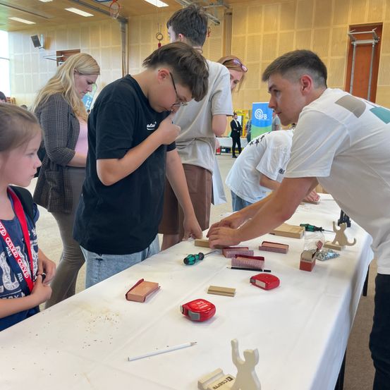 Eine Gruppe von Menschen, einschließlich Erwachsener und Kinder, sind um einen Tisch versammelt und beteiligen sich an einem Workshop oder einer Aktivität. Sie verwenden Werkzeuge und Materialien.