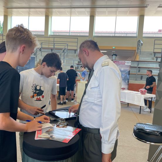 Drei junge Männer stehen an einem Tisch mit einem Mann in Uniform. Sie betrachten ein Tablet auf dem Tisch, und einer schreibt. Dahinter steht ein Tisch mit einer weißen Tischdecke und einer Schüssel mit Obst. In der Ferne sitzen Menschen auf Tribünen.