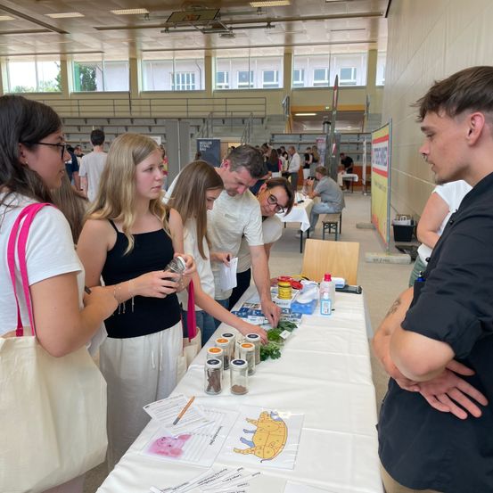 Mehrere Menschen, darunter Frauen und Männer, sind um einen Tisch mit Gläsern und einem Bleistift versammelt. Sie scheinen an einer Veranstaltung oder einem Workshop teilzunehmen. Ein Mann in Schwarz steht in der Nähe.