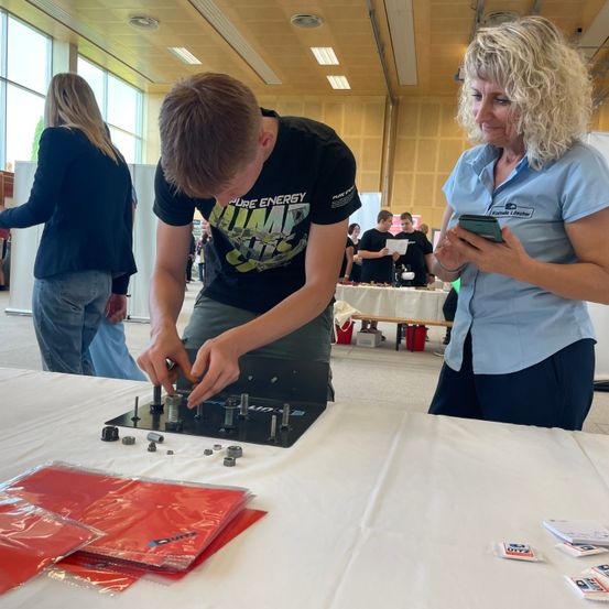 Ein junger Mann baut Komponenten auf einer schwarzen Tafel in einer Werkstatt zusammen. Eine ältere Frau beobachtet und hält ein Tablet. In der Nähe stehen Menschen hinter einem Tisch, und eine Glaswand ist im Hintergrund zu sehen.
