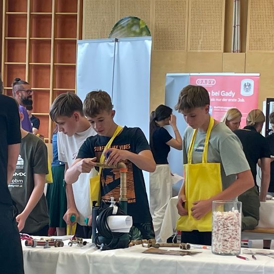 Eine Gruppe junger Jungen in gelben Schürzen steht um einen Tisch herum und arbeitet möglicherweise an einem Projekt. Andere Leute sind im Hintergrund, einige schauen sich etwas am Tisch an. Eine rosafarbene Fahne ist an der Wand.