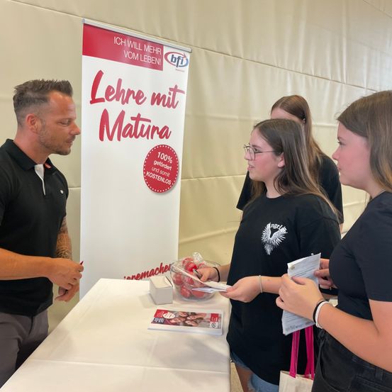 Ein Mann und zwei Frauen stehen an einem Tisch und sehen sich an. Der Mann hält einen Stift, und die Frauen halten Papiere. Ein Banner hinter ihnen lautet 'Lehr mit Matura'.