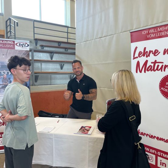 Ein Mann und zwei Frauen stehen an einem Tisch, wahrscheinlich auf einer Jobmesse. Der Mann gibt einen Daumen nach oben. Ein Banner hinter ihnen lautet Lehre mit Maturität.