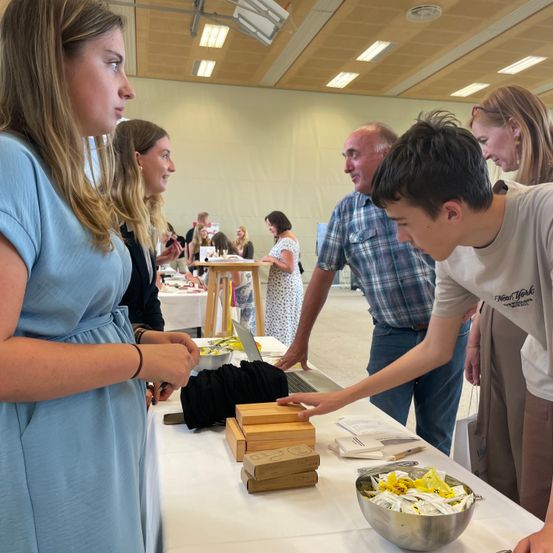 Eine Gruppe von Menschen untersucht Produkte an einem Tisch. Ein junger Mann zeigt auf Artikel. Frauen in Kleidern und ein Mann in einem karierten Hemd schauen zu. Eine Schüssel mit Produkten steht auf dem Tisch.