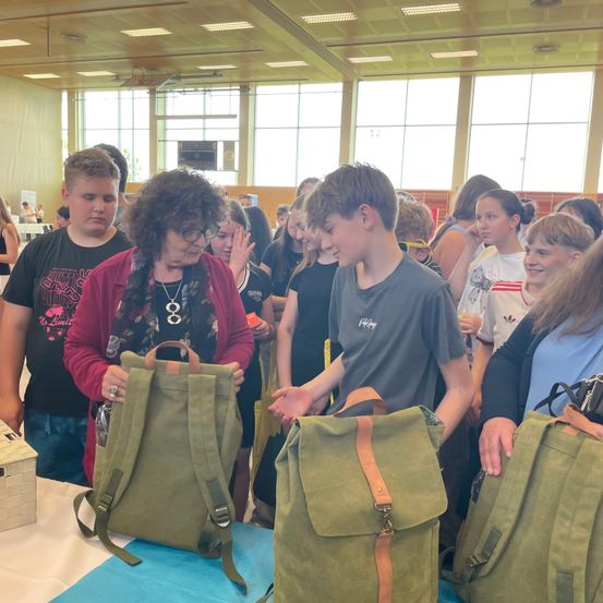 Eine Gruppe von Menschen in einem Saal betrachtet grüne Rucksäcke. Eine Frau hält einen und ein Junge neben ihr untersucht ihn. Ein anderer Junge vor ihnen hält einen Rucksack. Es stehen weitere Menschen hinter ihnen.