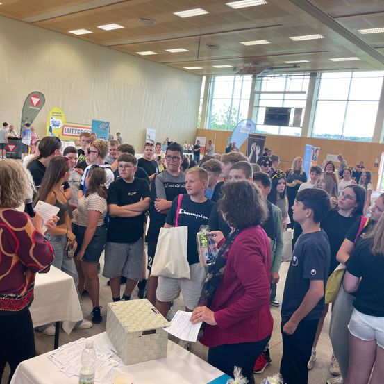 Eine Gruppe von Menschen versammelt sich drinnen, einige stehen, andere warten, während eine Frau an einem Tisch Gegenstände verteilt. Banner und Poster hängen an den Wänden.