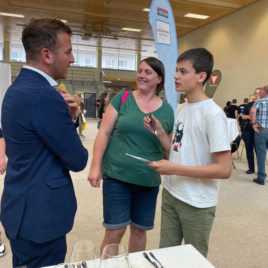 Ein Mann und eine Frau unterhalten sich mit einem Jungen vor einem Tisch. Der Mann trägt einen blauen Anzug, die Frau ein grünes T-Shirt und der Junge ein weißes T-Shirt. Hinter ihnen gehen Menschen in einem großen offenen Raum.