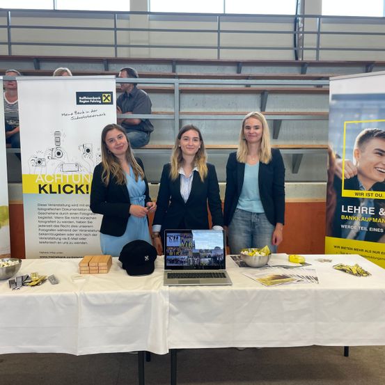 Drei Frauen stehen an einem Tisch mit Laptop und Werbematerialien. Hinter ihnen werben Plakate für eine Banklehre. Zuschauer sitzen auf den Tribünen.